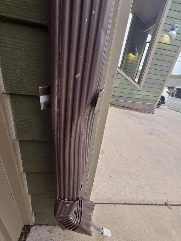 A brown downspout with a dented lower section beside a concrete walkway